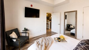 A hotel room with a bed, a chair, a small table with a plant, a TV mounted on the wall, and a mirror reflecting the room.