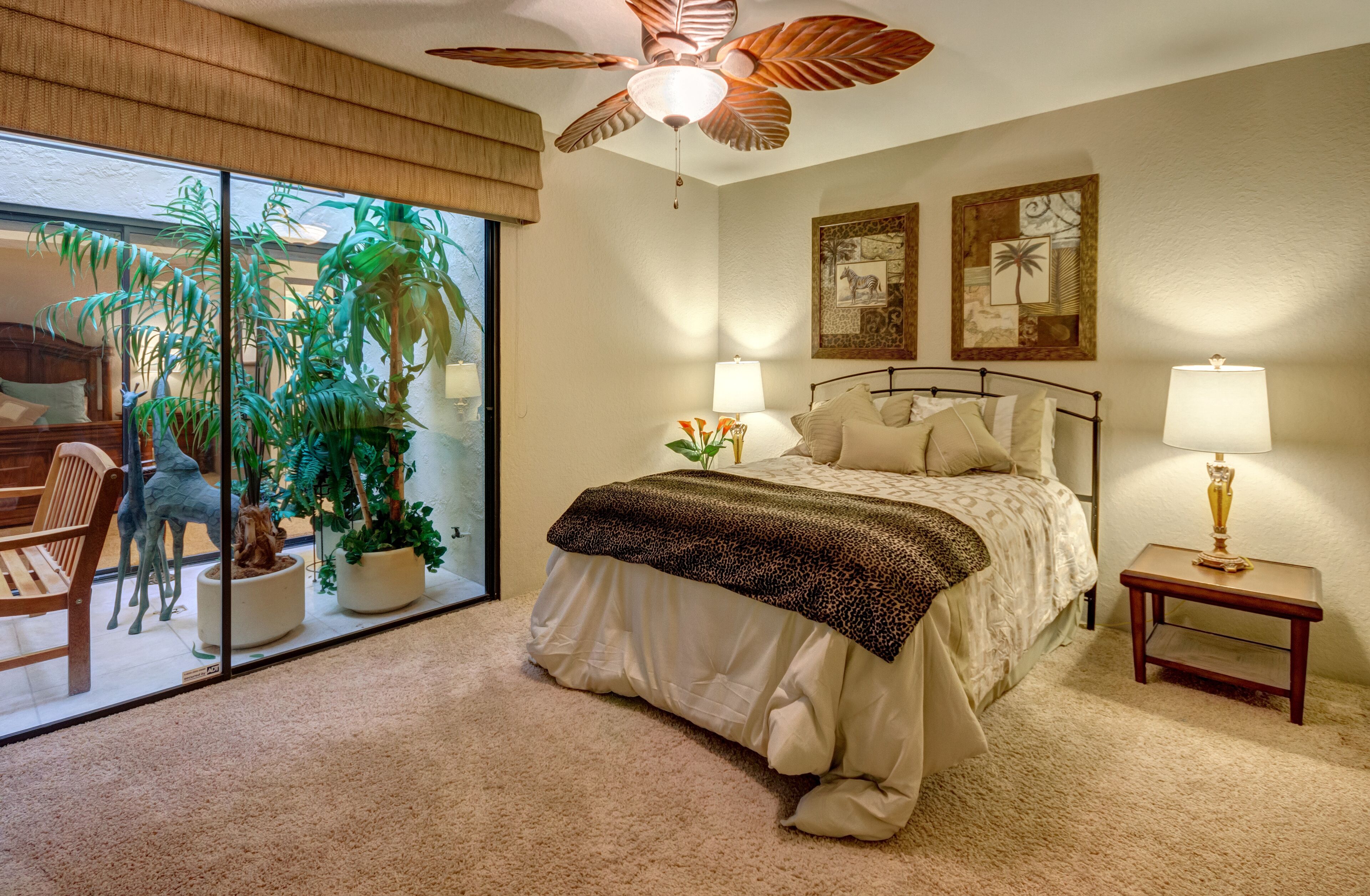 A bedroom with a bed, two bedside tables with lamps, a ceiling fan, and a large window with a view of a patio area.