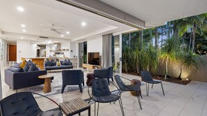 Ein moderner Wohnbereich mit Sofa, Sesseln und einem Essbereich. Im Hintergrund befindet sich eine Küche und es gibt einen Blick auf tropische Pflanzen vor den Fenstern.