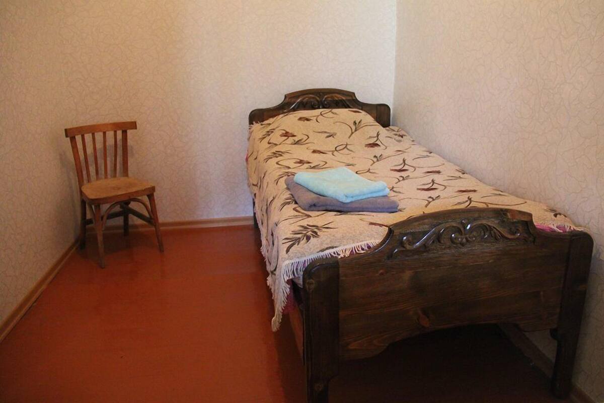 A small, single-bed room with a wooden headboard and a floral-patterned bedspread. A wooden chair is positioned against the wall.
