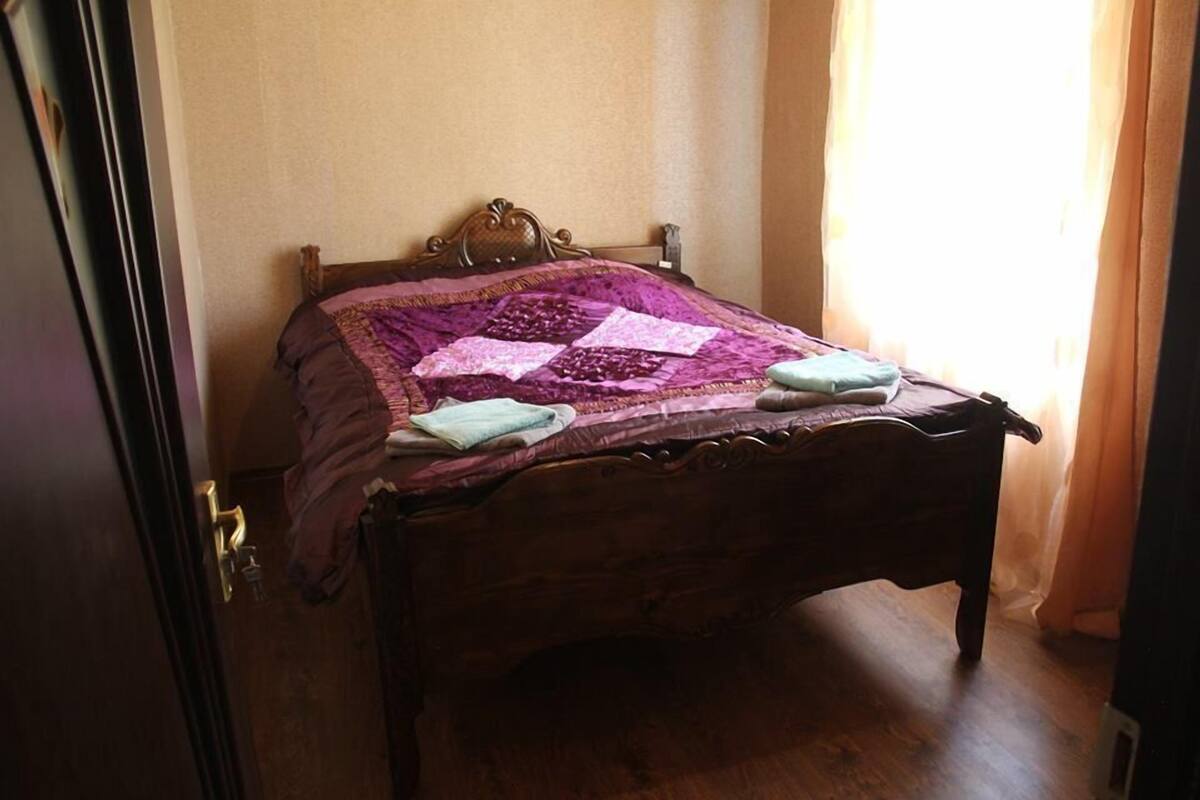 A wooden bed with a purple quilt, a nightstand, and a window with curtains.