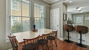 A dining area with a wooden table and four chairs, two bar stools, and large windows with blinds.