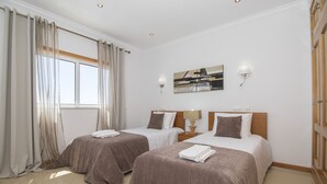 A hotel room with two beds, a window with curtains, a painting on the wall, and a nightstand with a lamp.