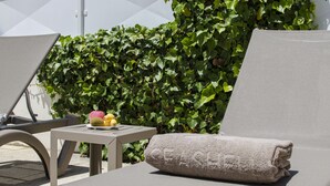 A sun lounger with a towel, a small table with fruit, and a hedge in the background.