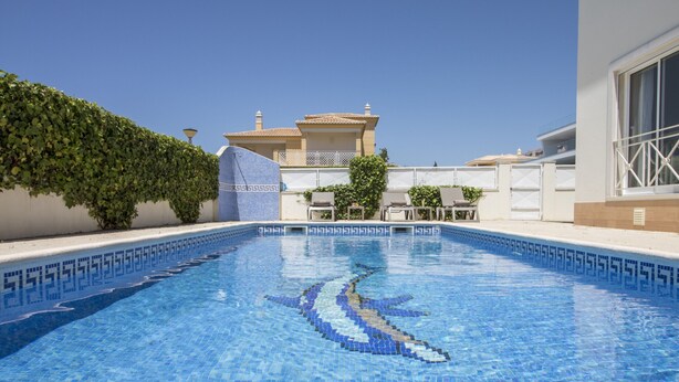 A swimming pool with a mosaic design, a white building with a balcony, and a hedge.