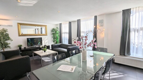 Una sala de estar moderna con una mesa de comedor de vidrio, sofás negros, un televisor de pantalla plana y un espejo grande.