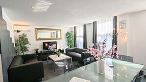 Una sala moderna con un sofá, una mesa de centro, un televisor y una mesa de comedor con sillas. Hay una planta y un florero con flores en la mesa.