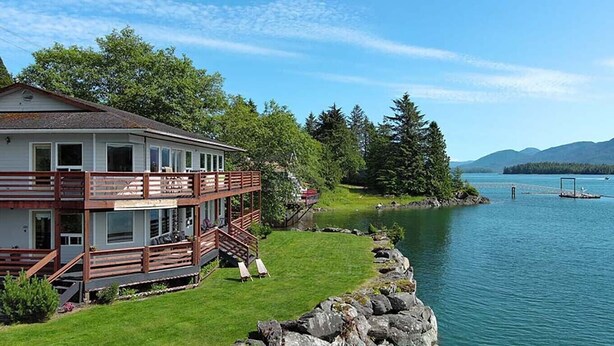 A two-story house with a balcony overlooking a lake, surrounded by greenery and mountains in the background. - Waterfront only 2 miles from downtown!  Sleeps 8. Amazing Views! (Ketchikan)