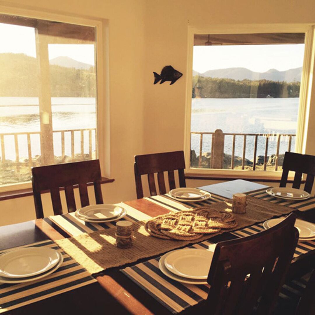Un coin repas offrant une vue sur un lac et des montagnes, avec des chaises en bois, une nappe à rayures et une décoration représentant un poisson sur le mur.