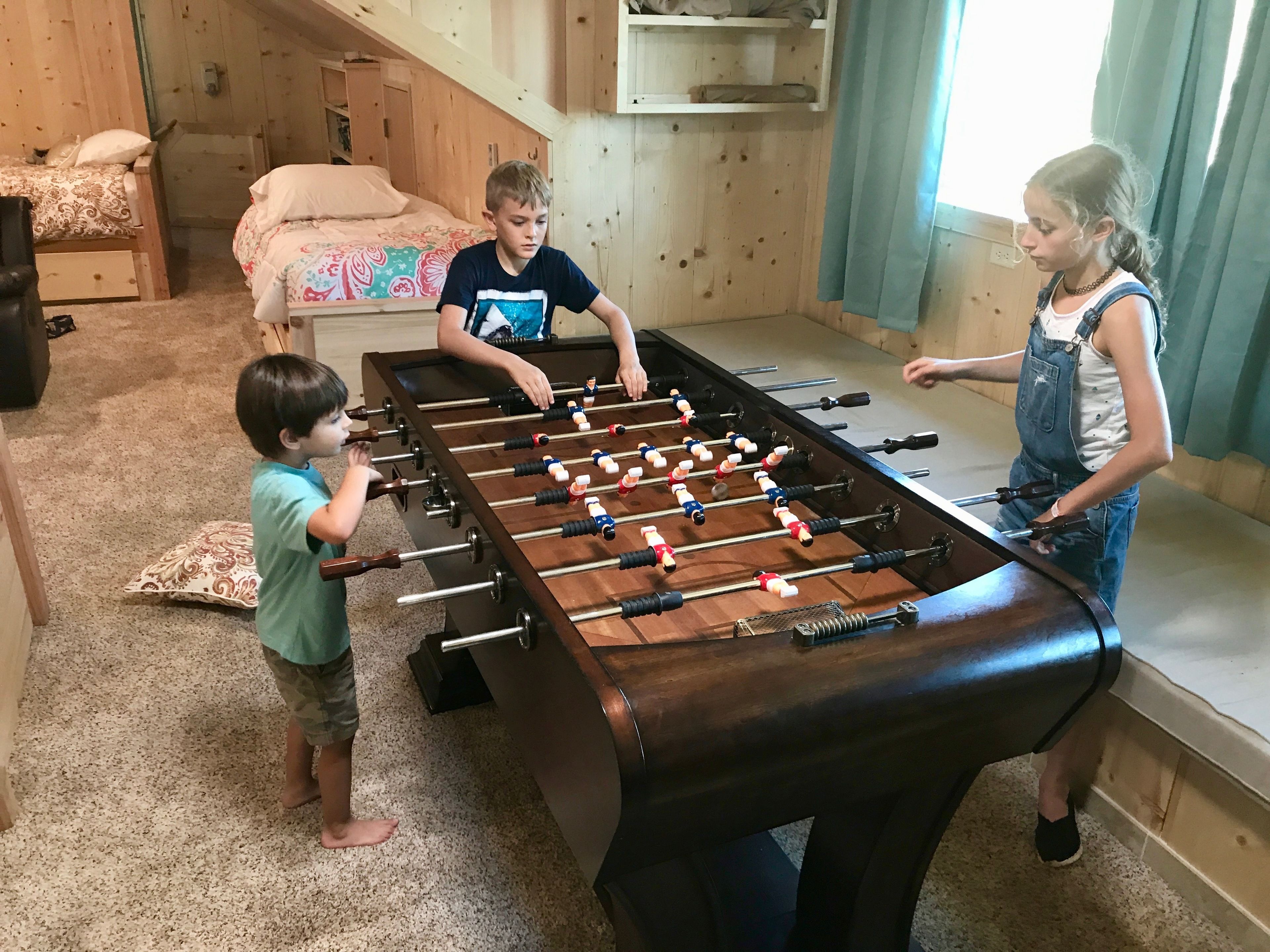 Une pièce avec une table de baby-foot, un lit recouvert d’une couvre-lit à motifs et une fenêtre avec des rideaux.