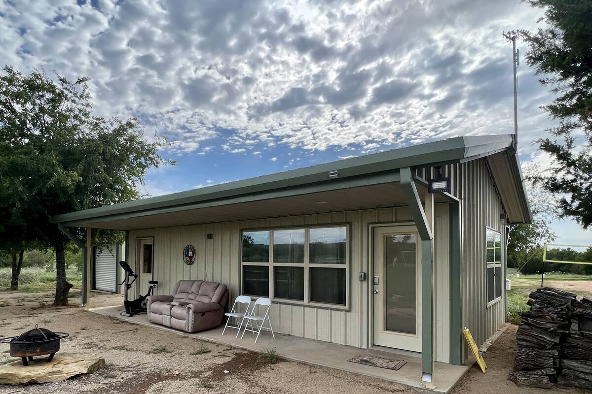 A small, single-story building with a covered porch, a sliding glass door, and a patio area with a sofa and chairs.
