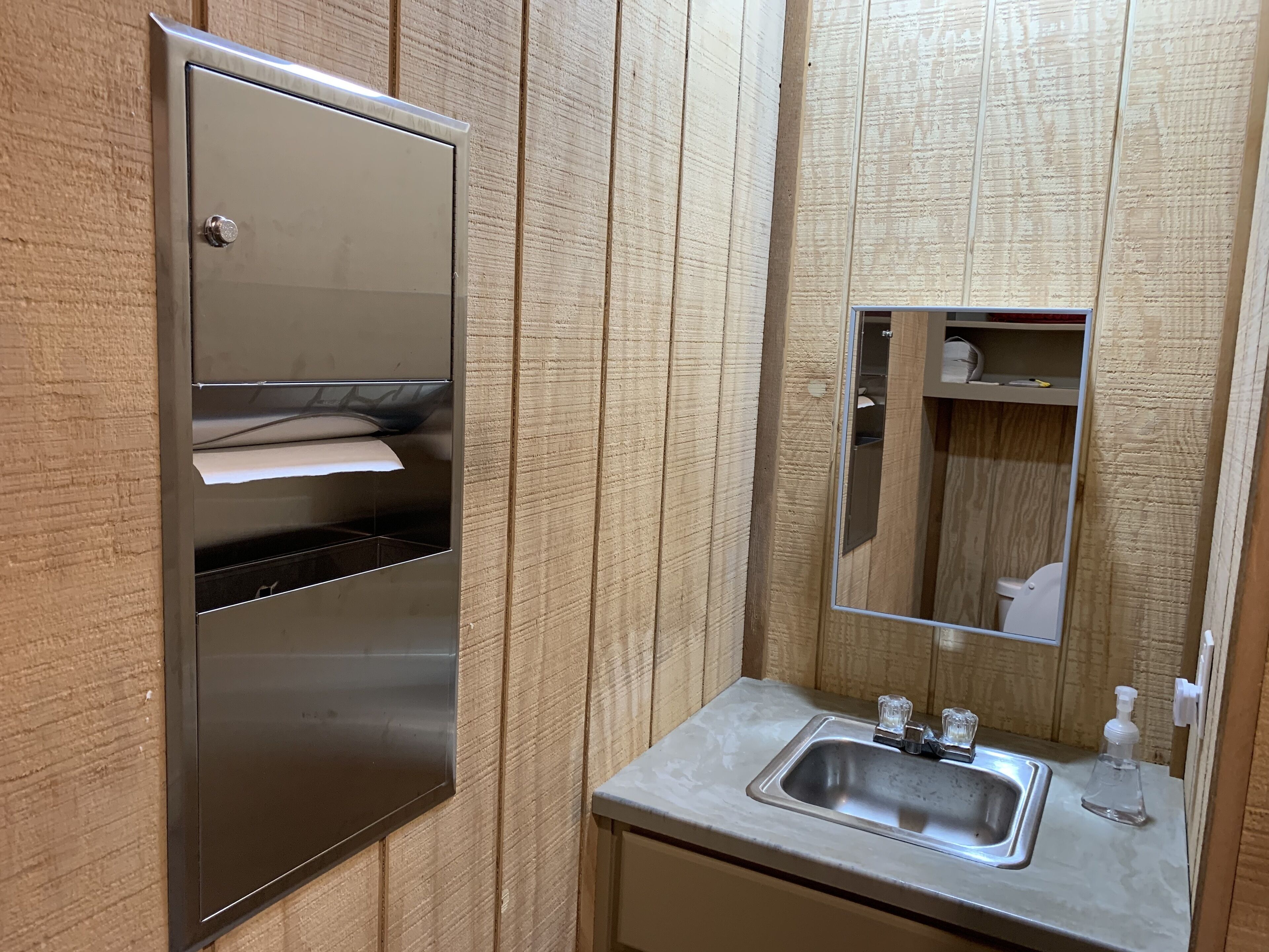 A bathroom with a stainless steel towel dispenser, a sink with a mirror, and a wooden panel wall.