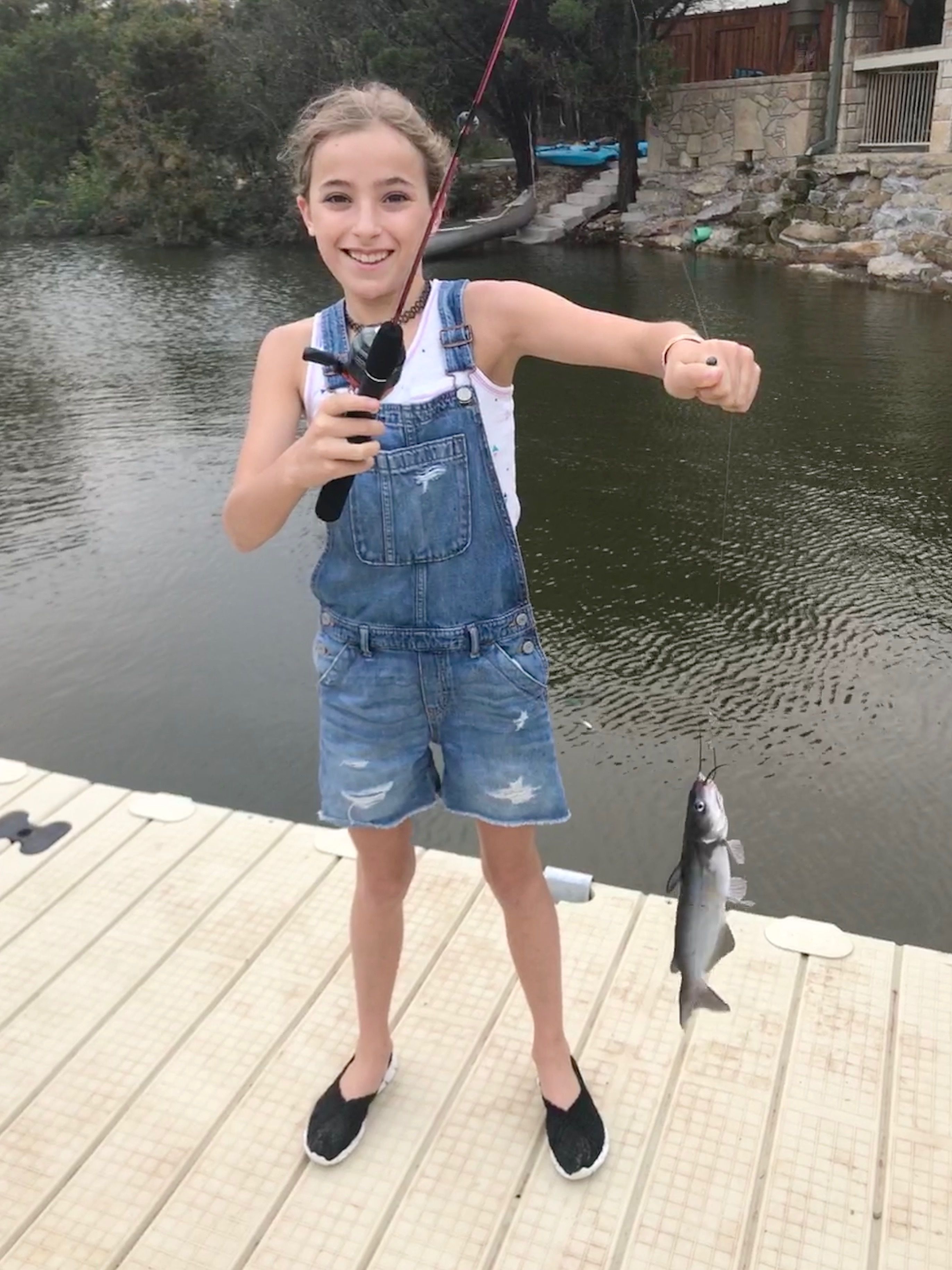 Une personne se tient sur un quai, tenant un poisson, et porte une salopette en jean et des chaussures noires.