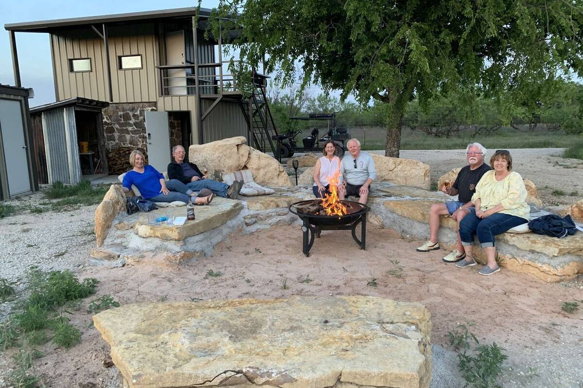 A group of people sitting around a stone fire pit in a rustic outdoor setting with a stone building in the background.