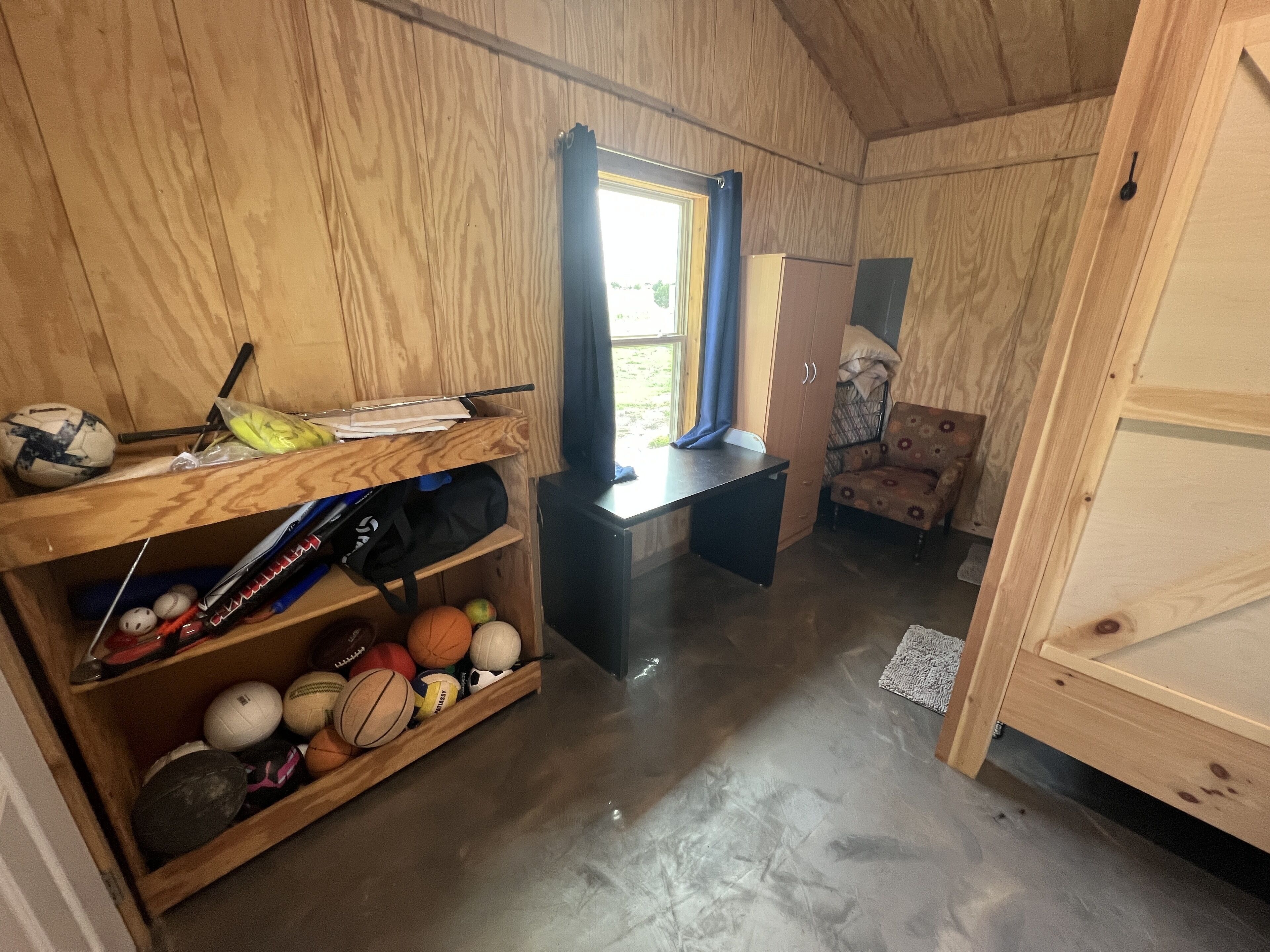 Una habitación de madera con un escritorio, estantes llenos de balones deportivos, una silla y un armario.