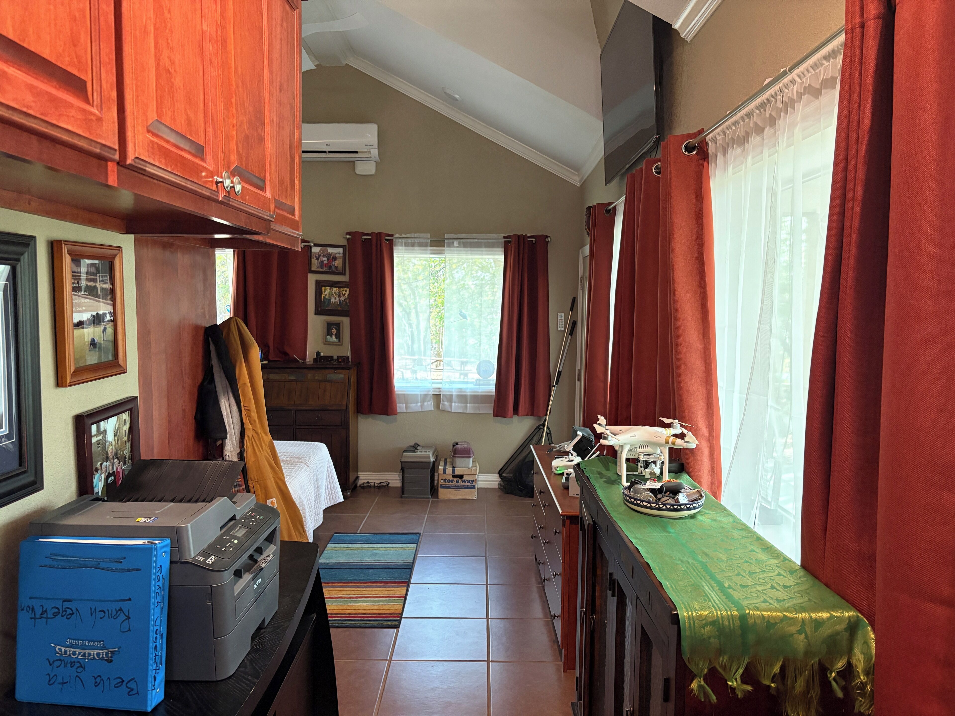 A compact, well-lit room with wooden cabinets, a bed, a desk with a printer, a window with curtains, and a small table with a drone model.