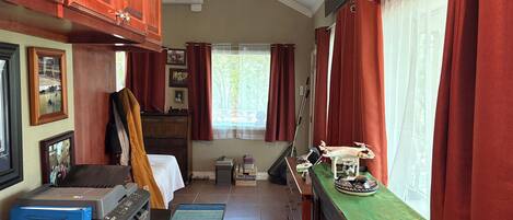 Una camera compatta e ben illuminata con mobili in legno, un letto, una scrivania con una stampante, una finestra con tende e un piccolo tavolo con un modellino di drone.