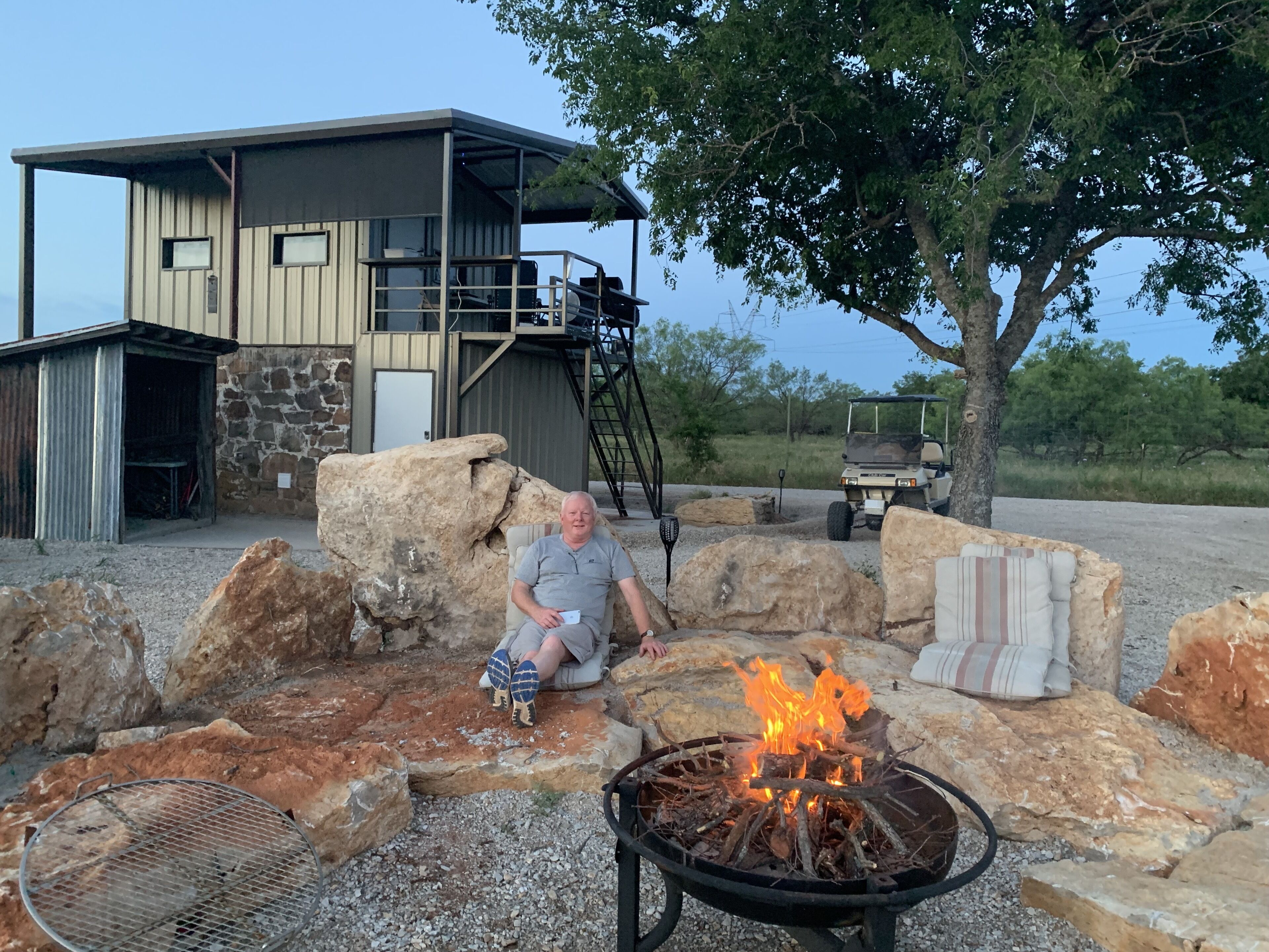 A man sitting by a fire pit with a stone building in the background.