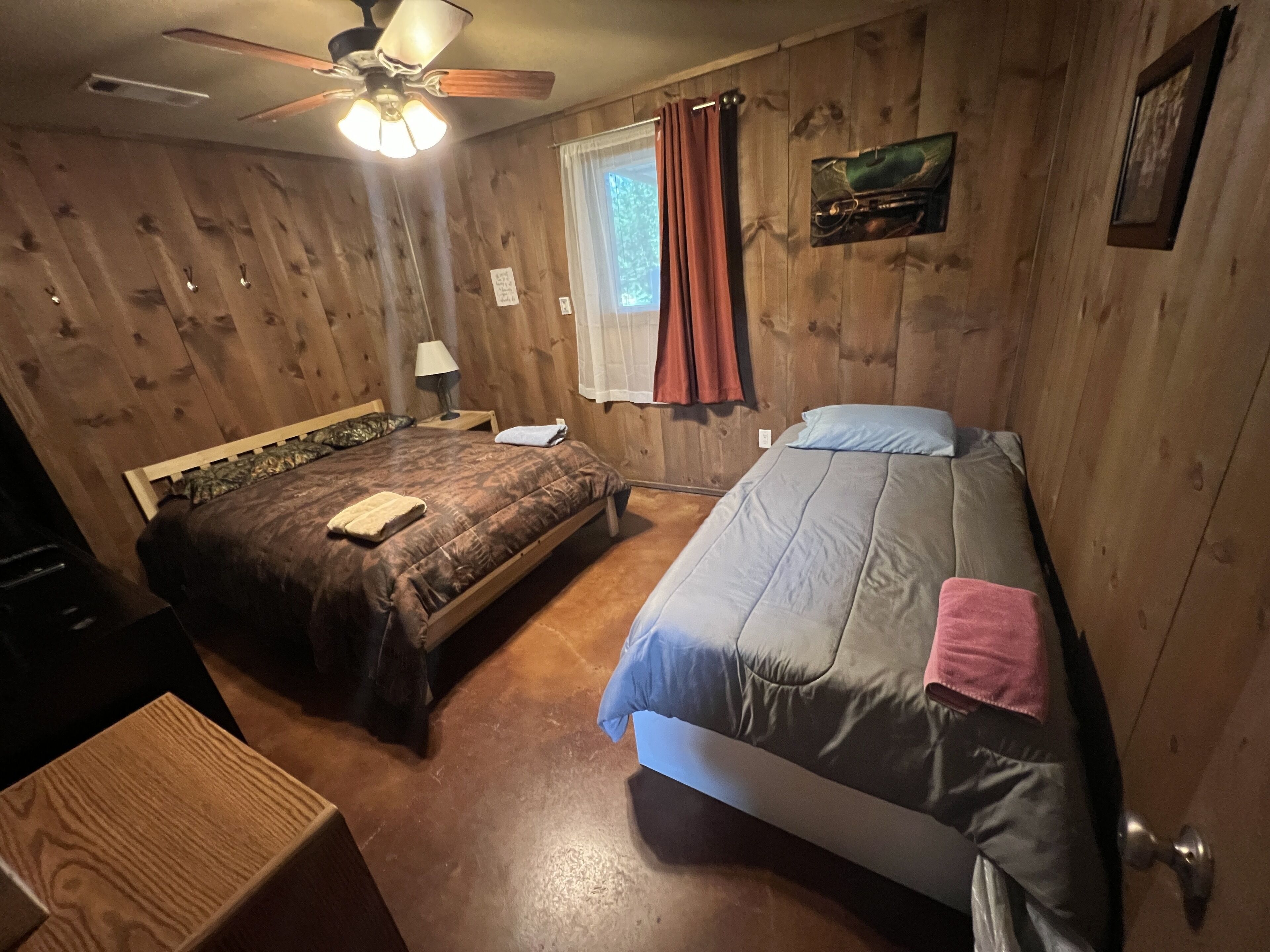 Quarto com duas camas, paredes de madeira, ventoinha no teto, janela com cortinas e uma pequena mesa com abajur.