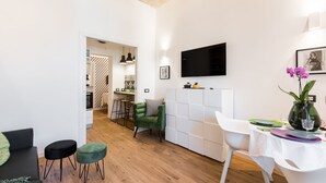 A modern living room with a sofa, two ottomans, a dining table with chairs, a TV mounted on the wall, and a kitchen area in the background. - Charming Trastevere home overlooking a medieval church: suite, 3 baths, WiFi, AC (Roma)