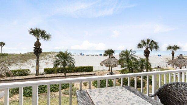 A balcony with a view of the beach, palm trees, and a thatched umbrella.