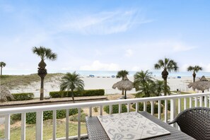 A balcony with a view of the beach, palm trees, and a thatched umbrella.
