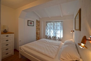 A bedroom with a bed, a dresser, a window with floral curtains, and a wall-mounted lamp.