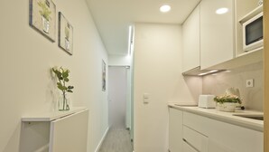 A narrow hallway leading to a kitchen with white cabinets, a microwave, and a toaster. There are framed pictures on the wall and a small plant on the counter. - OLD TOWN PRÍNCIPE REAL - Apart. modern with patio and direct access to quiet street. (Lisboa)