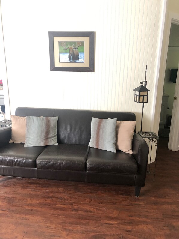 A dark brown leather sofa with three cushions, a framed picture of a moose on the wall, and a floor lamp.
