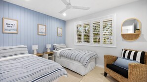 Ein Schlafzimmer mit zwei Betten, einem Deckenventilator, einem Fenster mit Rollläden und einem Korbstuhl.
