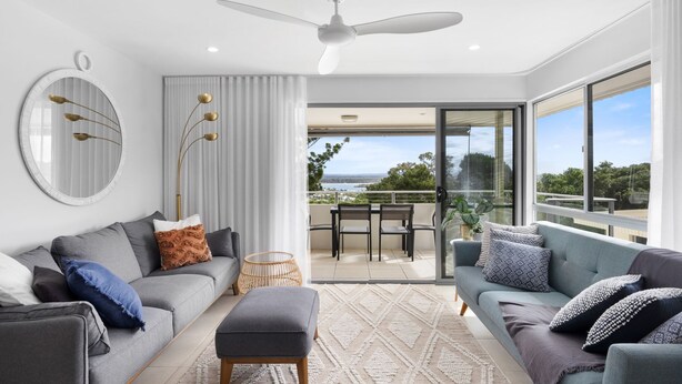 Ein modernes Wohnzimmer mit grauem Sofa, blauem Sofa und Essbereich mit Tisch und Stühlen. Ein großes Fenster bietet Blick ins Freie.