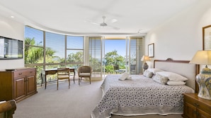 A bedroom with a large bed, a desk, and a chair. There is a TV on the wall and a window with blinds.