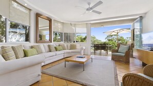 A modern living room with a white sectional sofa, a coffee table, and a TV. There is a balcony with a table and chairs, and a ceiling fan.