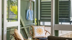 A room with a view of a green fence, a wicker chair with a sun pillow, and a hanging bag with a blue strap.