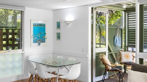 A modern dining area with a glass table and white chairs, a framed painting, and a view of greenery through the glass door.