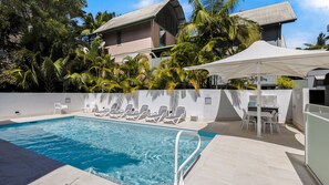 A swimming pool with lounge chairs and a barbecue area, surrounded by palm trees and a building with a sloped roof.
