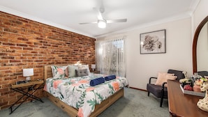 A bedroom with a brick wall, a bed with floral bedding, a ceiling fan, a bedside table with a lamp, and a chair with a vase of flowers. - 29A Ballina Crescent, Port Macquarie (Port Macquarie)