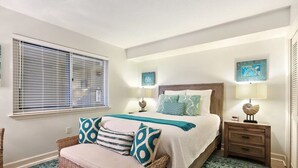 A bedroom with a bed, a nightstand, a wicker chair, a window with blinds, and two wall-mounted lamps. - Sandy Key 635: 2 BD/2BH, 6th floor (Pensacola)