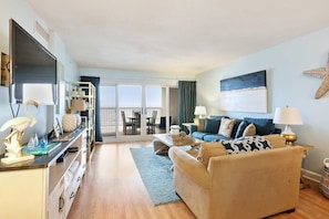 A spacious living room with a sofa, coffee table, and TV. There is a balcony with a view.