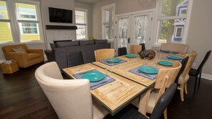 Un coin repas avec une table en bois dressée pour six personnes, entourée de chaises, dans une pièce où se trouvent un canapé et un téléviseur fixé au mur.