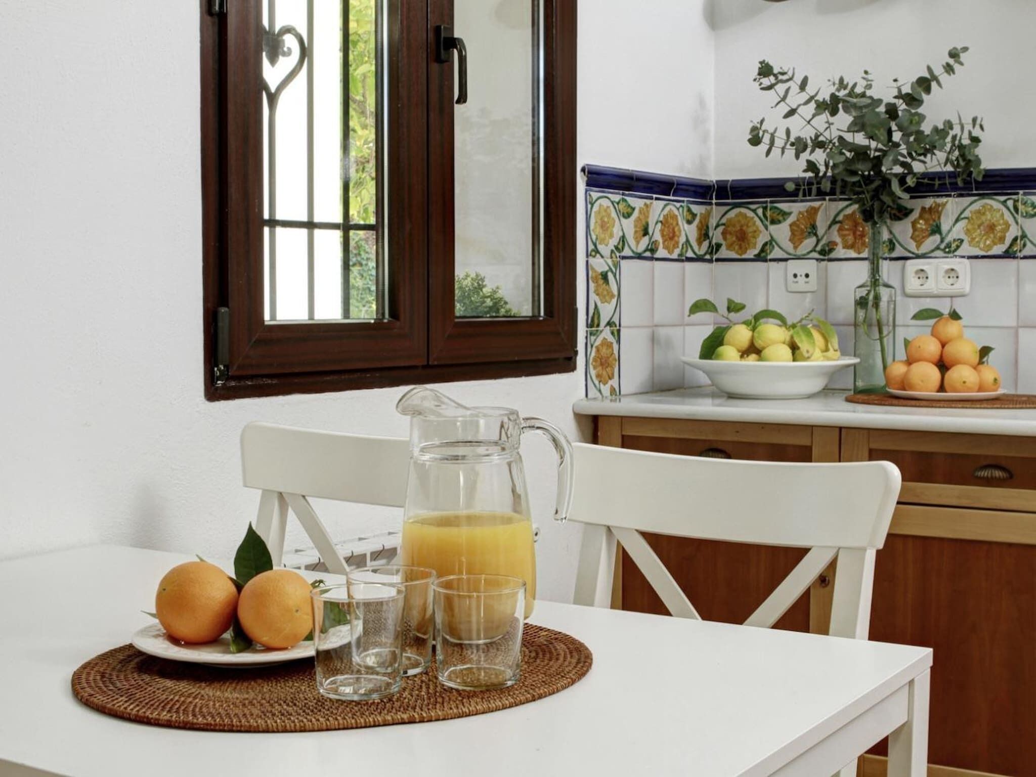 A kitchen with a white table set for breakfast, including oranges, a pitcher of orange juice, and glasses.