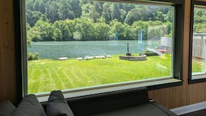 Ein Zimmer mit großem Fenster mit Seeblick, eine kleine Terrasse mit Stühlen und ein Steinbau.