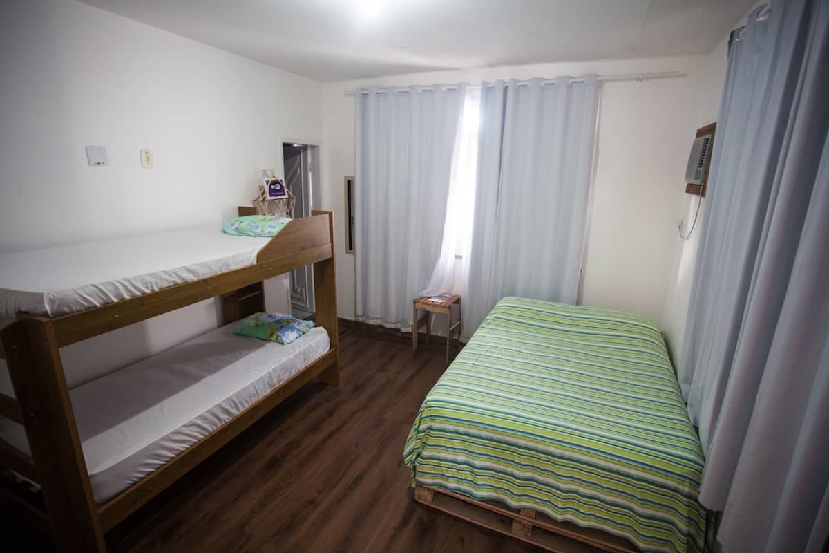 A room with a bunk bed, a single bed, a small table, and a window with curtains.