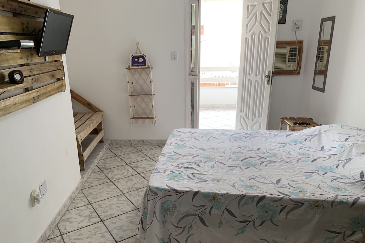 A bedroom with a bed, a TV mounted on a wooden shelf, a WiFi sign, a small table, and a window with a view.