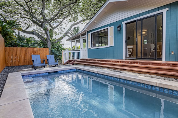 Une piscine avec une terrasse en bois, un patio couvert et une vue sur une maison dotée de grandes fenêtres et d’une porte coulissante.