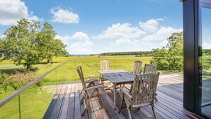 Une terrasse en bois avec une table et des chaises, donnant sur un champ verdoyant et des collines lointaines.