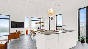 Une cuisine moderne dotée d’un grand îlot central, d’un coin repas avec des chaises, d’un téléviseur fixé au mur et de grandes fenêtres offrant une vue dégagée.