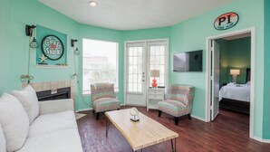 A room with teal walls, a white sofa, two patterned chairs, a wooden coffee table, a fireplace, a television, and a bed with a white comforter. - Cozy Coastal Condo with Lake Views, Heated Pool, and Easy Beach Access (Corpus Christi)