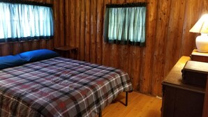 Una habitación con paredes de madera, una cama con colcha escocesa, dos ventanas con cortinas, una mesita y una lámpara en una cómoda.