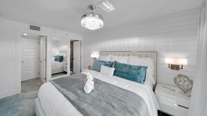 A hotel room with a large bed, a chandelier, a nightstand with a lamp, and a door leading to another room.